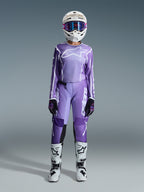 Pilota di motocross con completo Alpinestars Stella Fluid Apex 2026, maglia viola e bianca con pantaloni coordinati, casco bianco con occhiali rosa e stivali da fuoristrada bianchi e neri, kit da gara completo per motocross femminile.