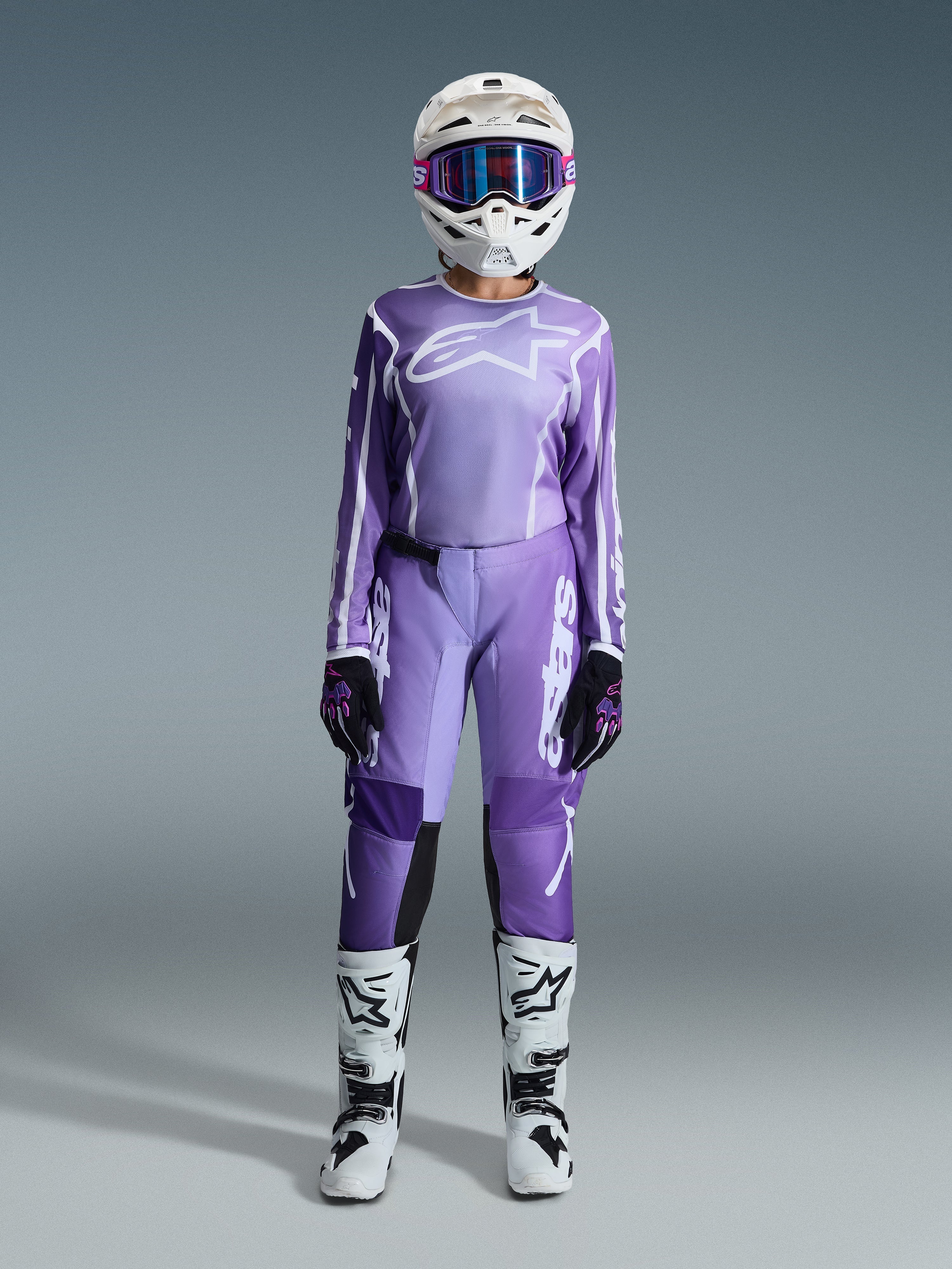 Pilota di motocross con completo Alpinestars Stella Fluid Apex 2026, maglia viola e bianca con pantaloni coordinati, casco bianco con occhiali rosa e stivali da fuoristrada bianchi e neri, kit da gara completo per motocross femminile.