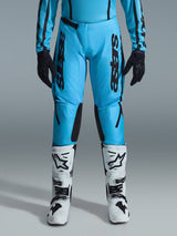 Alpinestars Stella Fluid Apex Pants 2026, pantaloni da motocross da donna, ciano brillante e nero, branding nero verticale sulle cosce, pannelli interni rinforzati neri sulle ginocchia, mostrati con stivali da motocross bianchi e neri e guanti neri, equipaggiamento professionale da fuoristrada per donna