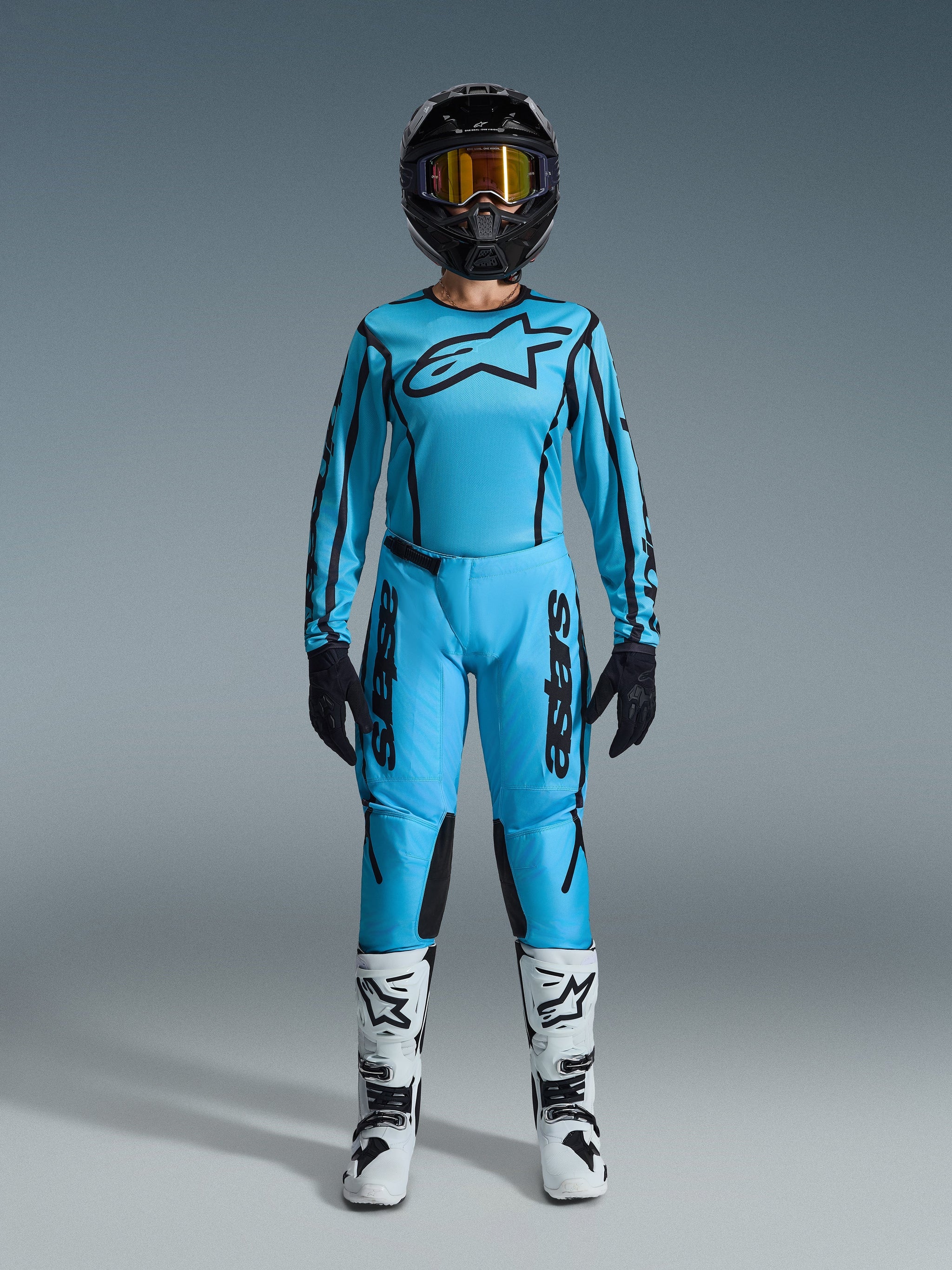 Pilota di motocross donna con set Alpinestars Stella Fluid Apex 2026, maglia e pantaloni azzurro ciano brillante con dettagli neri, casco nero con maschera iridescente e stivali bianchi, abbigliamento professionale completo da motocross