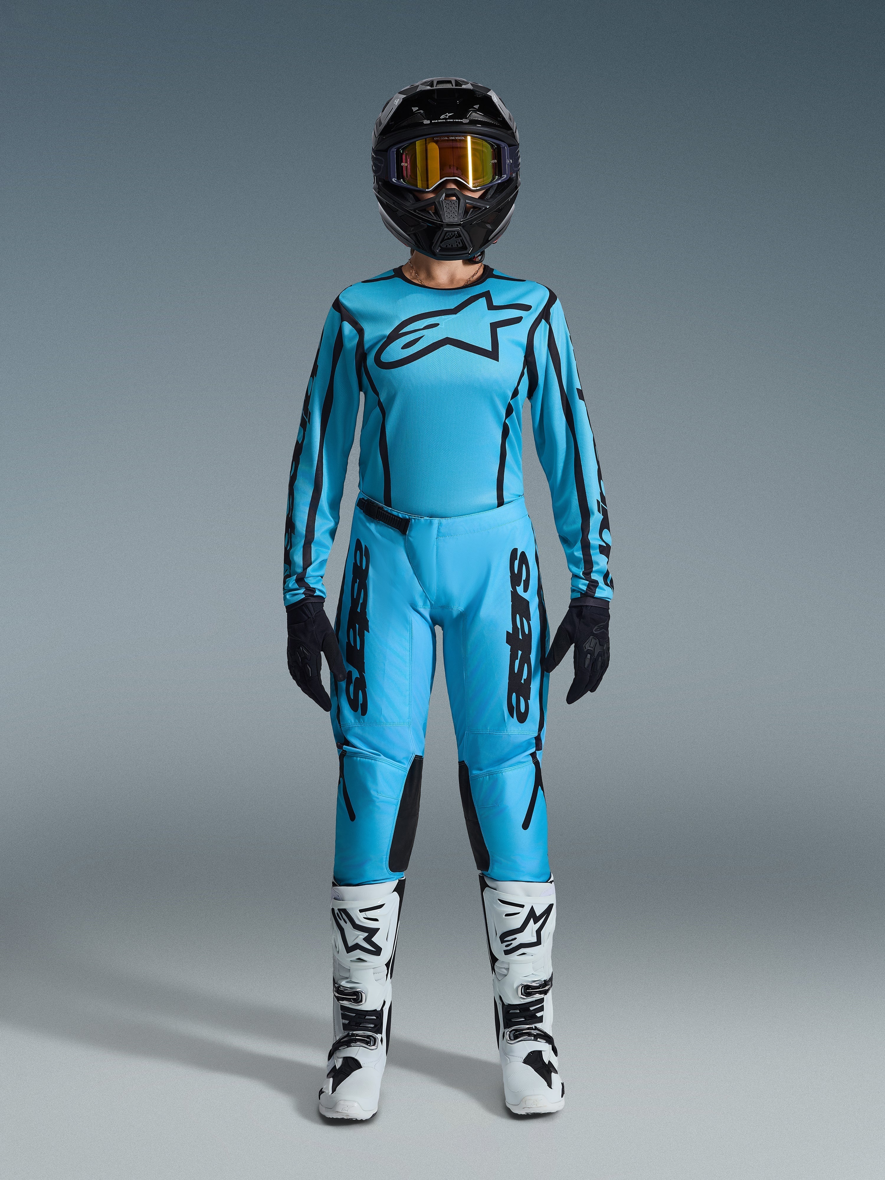 Alpinestars Stella Fluid Apex Gear Set 2026, maglia e pantaloni da motocross da donna, blu e nero, con grande logo a stella sul petto, indossato con casco nero, occhiali con lenti colorate e stivali bianchi con dettagli neri, completo da gara fuoristrada