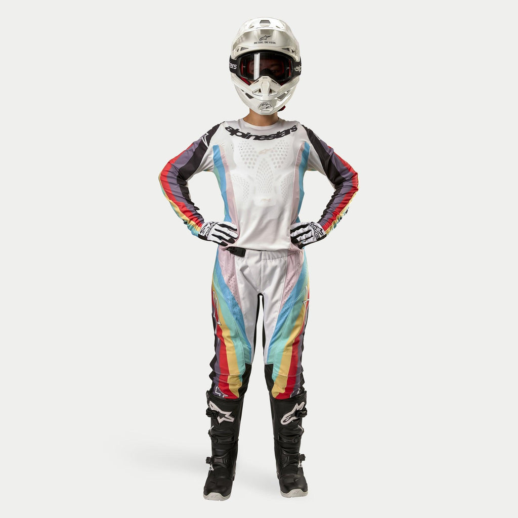 Pilota donna di motocross con pantaloni Alpinestars Stella Techstar 2024, Black Multicolor con base bianca e strisce rosse, gialle e blu, maglia a maniche lunghe coordinata, casco bianco e stivali neri, equipaggiamento da gara completo per fuoristrada