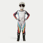 Pilota donna di motocross con pantaloni Alpinestars Stella Techstar 2024, Black Multicolor con base bianca e strisce rosse, gialle e blu, maglia a maniche lunghe coordinata, casco bianco e stivali neri, equipaggiamento da gara completo per fuoristrada