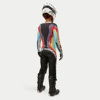 Pilota di motocross vista da dietro che indossa pantaloni Alpinestars Stella Techstar 2024 in Black Multicolor, con grande logo bianco sulla seduta, coordinati con una maglia a righe colorate, casco bianco e stivali Tech 3 neri per gare off-road.