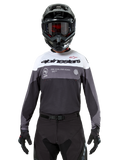 Pilota di motocross con maglia Alpinestars Pro-Dura in Black Magnet White, maglia tecnica a maniche lunghe con inserti bianchi e grigi, indossata con casco e occhiali neri, con logo 'alpinestars' e slogan 'ONE GOAL ONE VISION', abbigliamento da moto off-road