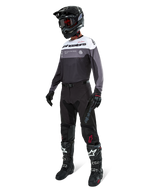 Maglia Alpinestars Pro-Dura in Black Magnet White, indossata da un pilota di motocross con equipaggiamento completo che include pantaloni neri, casco nero con occhiali e stivali bianchi e neri. La maglia presenta un design tecnico color-block con pannelli bianchi, grigi e neri, studiata per prestazioni enduro e off-road.