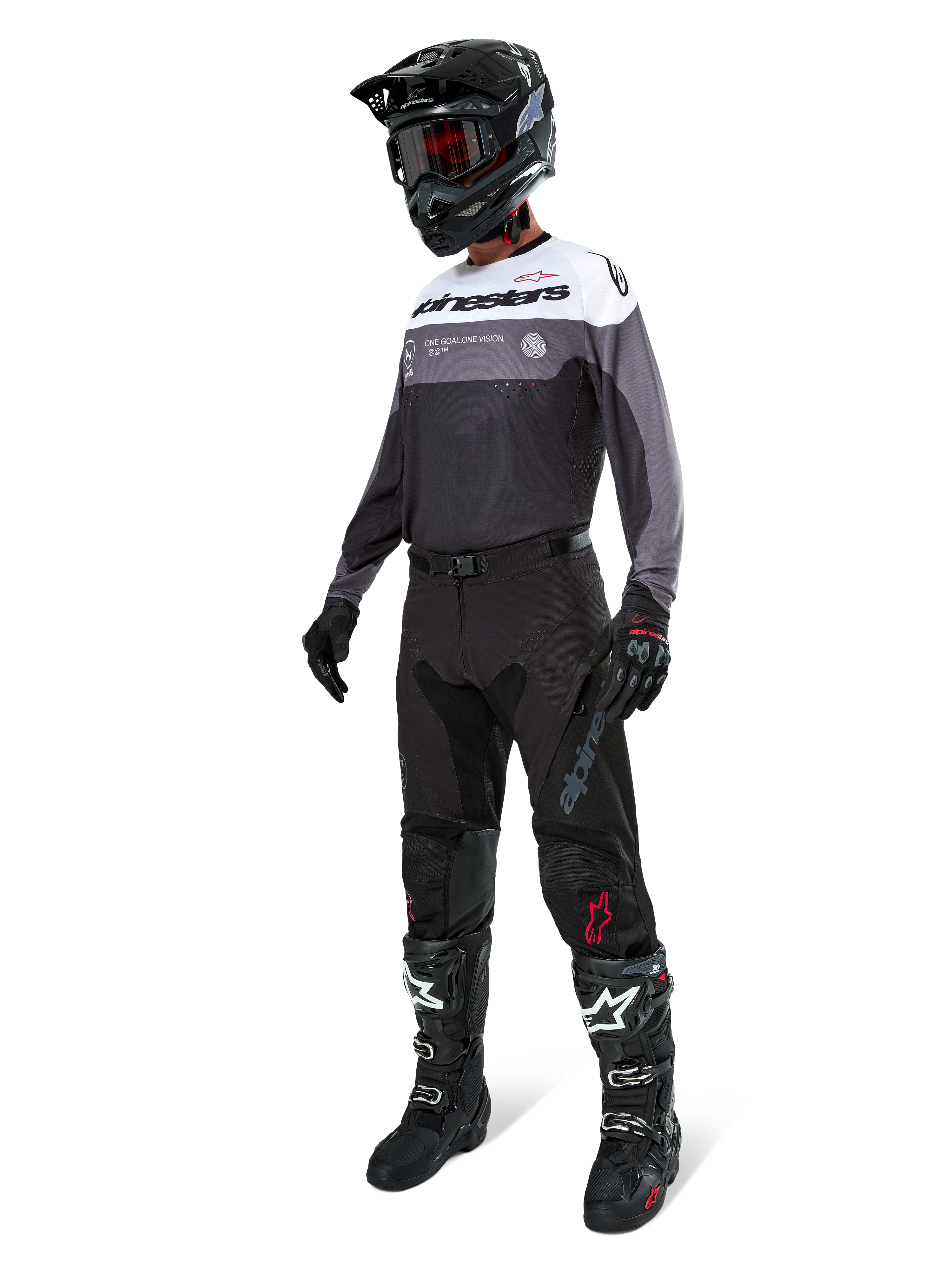 Maglia Alpinestars Pro-Dura in Black Magnet White, indossata da un pilota di motocross con equipaggiamento completo che include pantaloni neri, casco nero con occhiali e stivali bianchi e neri. La maglia presenta un design tecnico color-block con pannelli bianchi, grigi e neri, studiata per prestazioni enduro e off-road.