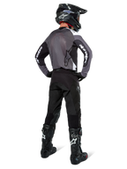 Pilota di motocross con maglia Alpinestars Pro-Dura in Black Magnet White, vista posteriore con pannello in mesh grigio e maniche nere con marchio Alpinestars bianco, coordinata con pantaloni neri, casco nero e stivali neri e bianchi, equipaggiamento professionale da enduro
