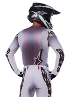 Alpinestars 2025 Supertech Maker Gear Set, pilota di motocross visto di schiena, maglia bianca e grigia con grande marchio nero sulle maniche e logo a stella, pantaloni bianchi coordinati con grafiche nere, casco bianco e nero, abbigliamento professionale da gara off-road
