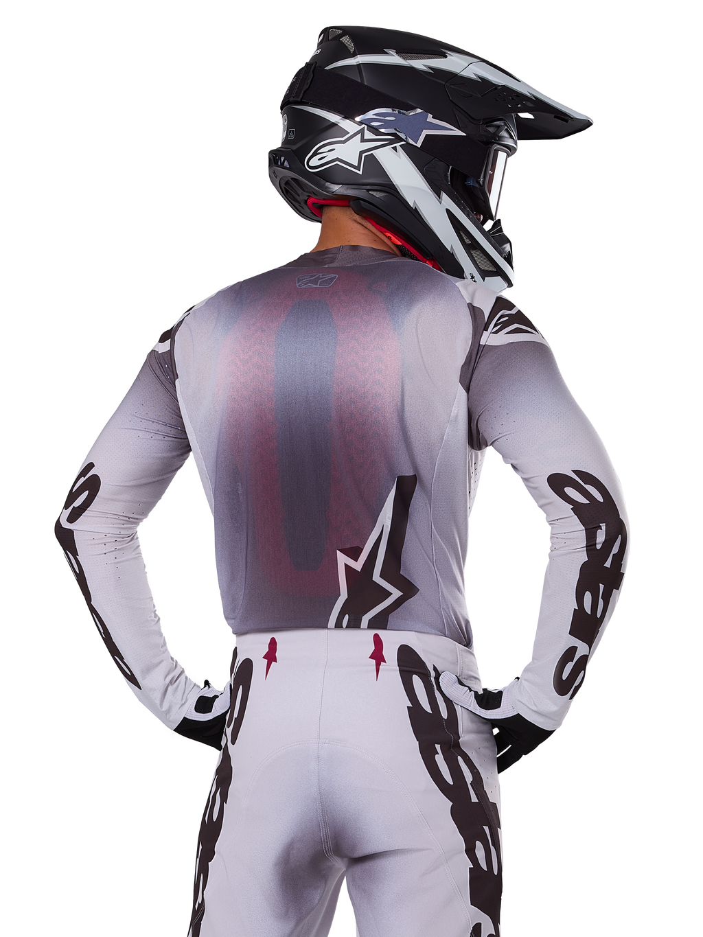 Pilota di motocross visto da dietro con indosso la maglia Alpinestars 2025 Supertech Maker, argento e nero con pannello posteriore in rete rossa e loghi neri sulle maniche, abbinata a pantaloni da motocross coordinati e casco bianco e nero, abbigliamento professionale per fuoristrada