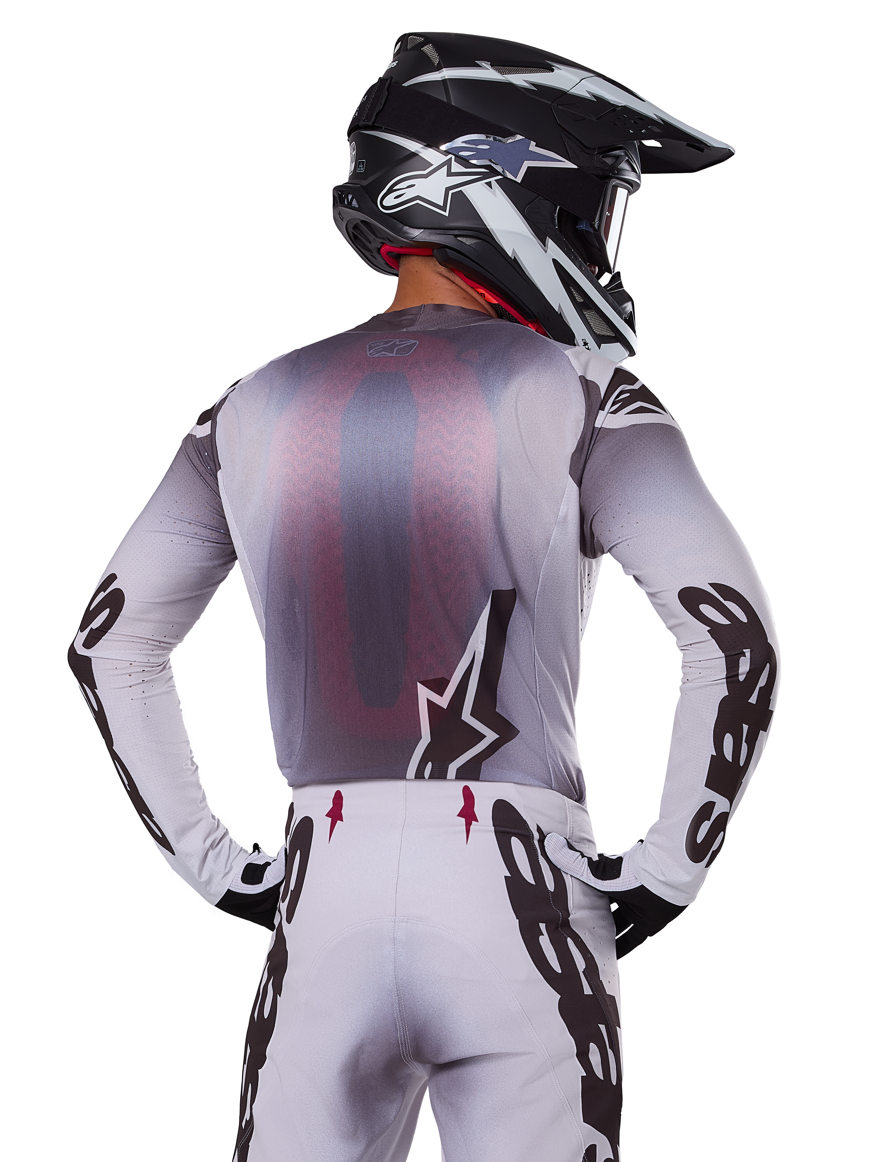 Pilota di motocross visto da dietro con indosso la maglia Alpinestars 2025 Supertech Maker, argento e nero con pannello posteriore in rete rossa e loghi neri sulle maniche, abbinata a pantaloni da motocross coordinati e casco bianco e nero, abbigliamento professionale per fuoristrada