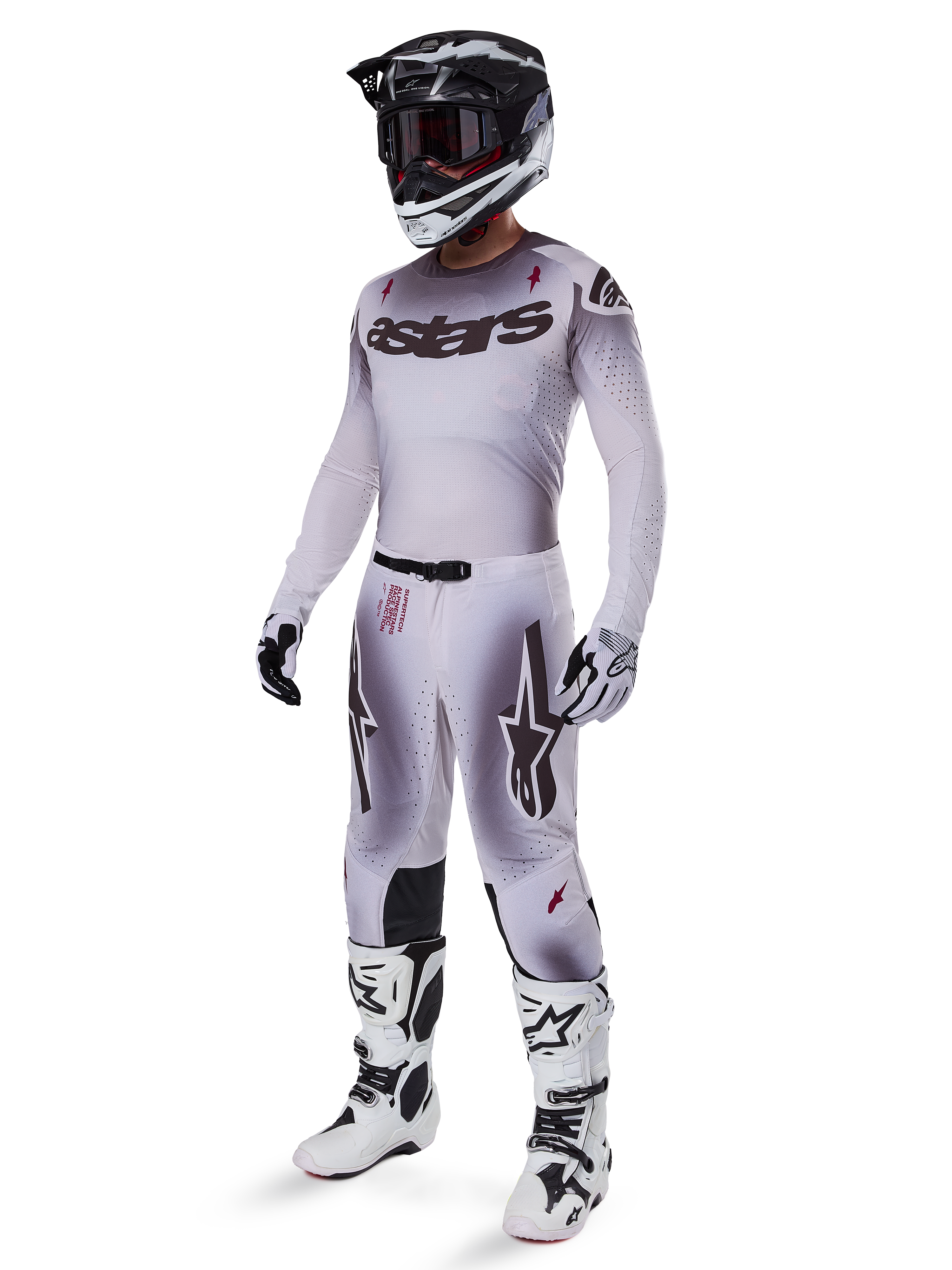 Pilota di motocross che indossa la maglia Alpinestars 2025 Supertech Maker in argento e nero, pantaloni coordinati argento e nero, casco bianco e nero con occhiali neri, stivali bianchi e neri e guanti coordinati, design professionale a gradiente per corse fuoristrada.
