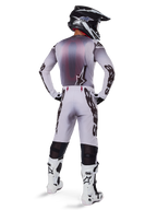 Pilota di motocross visto da dietro che indossa i pantaloni Alpinestars 2025 Supertech Maker in argento e nero, maglia a maniche lunghe coordinata, casco nero e bianco e stivali bianchi e neri, con evidenti loghi neri sulle gambe, abbigliamento professionale per gare fuoristrada.