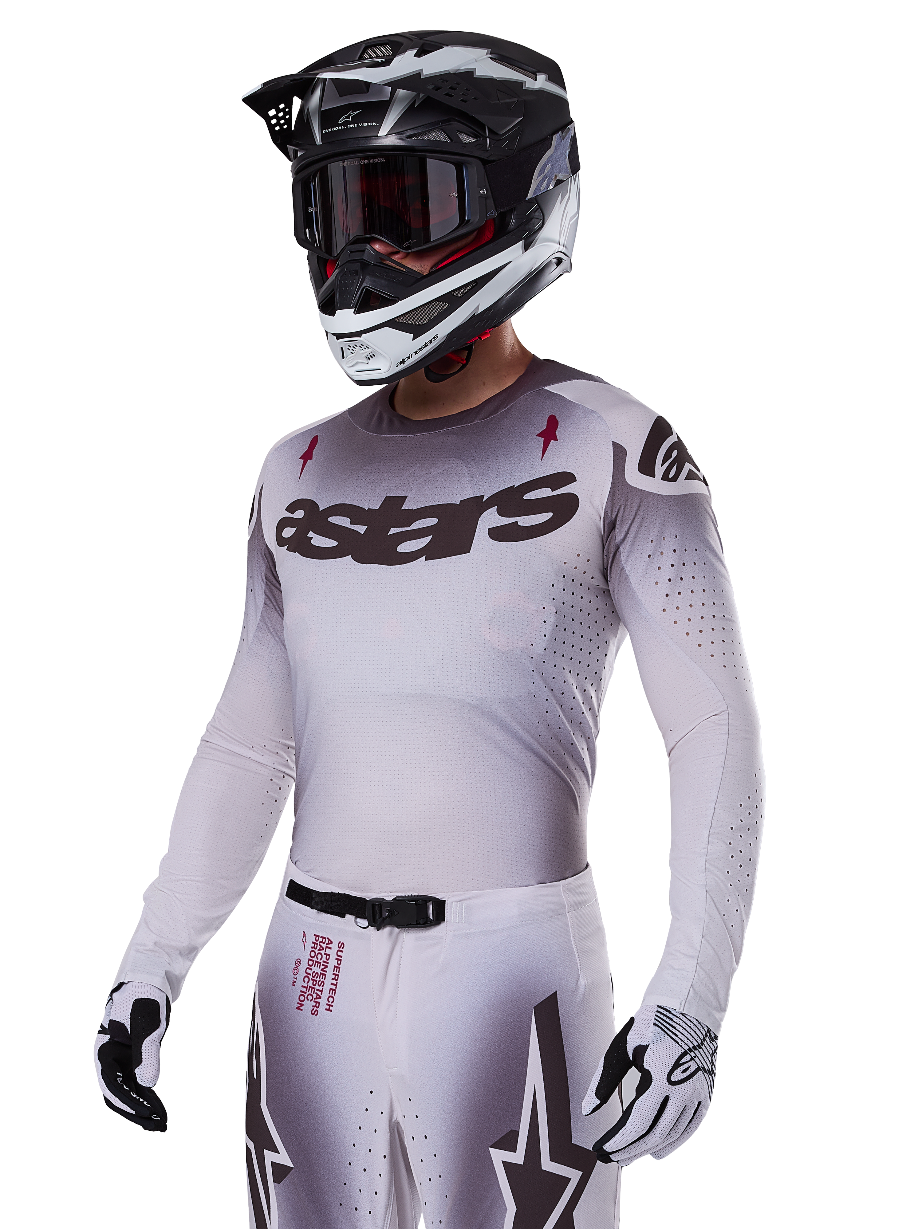 Pilota di motocross che indossa il set Alpinestars 2025 Supertech Maker in bianco, caratterizzato da una maglia con sfumatura grigia, logo astars nero e perforazioni tagliate al laser, pantaloni coordinati con fibbia magnetica e grafiche a stella, abbinato a casco e guanti bianchi e neri per un look completo da gara fuoristrada.