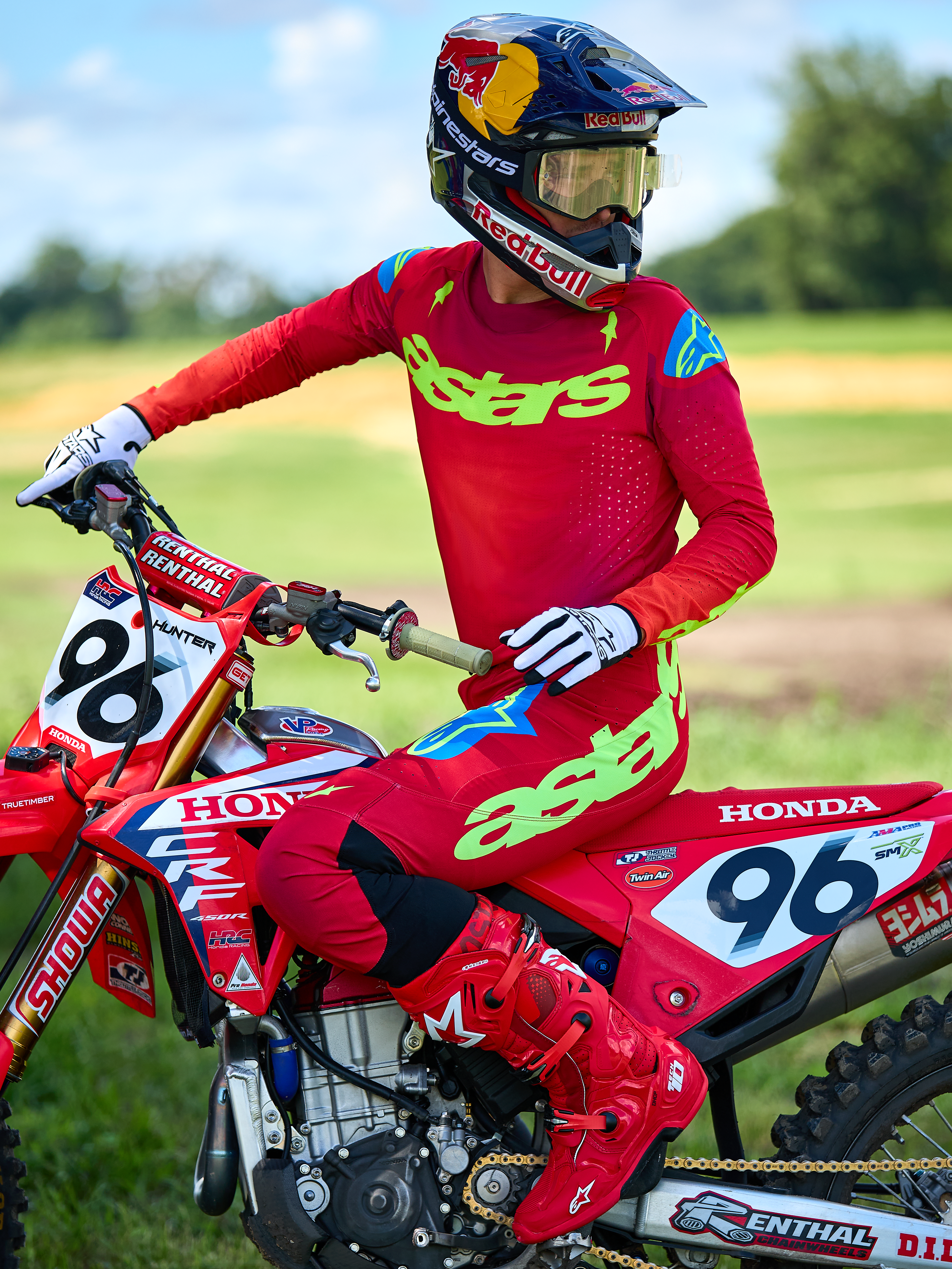 Pilota di motocross su moto Honda rossa con set Alpinestars 2025 Supertech Maker rosso, maglia con logo 'astars' verde neon e dettagli blu sulle spalle, pantaloni rossi coordinati, stivali rossi, guanti bianchi e casco Red Bull, equipaggiamento professionale per fuoristrada