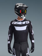 Alpinestars 2026 Racer Air Riway Jersey, maglia da motocross, nero e bianco con marchio 'astars', indossata da un pilota con casco nero, occhiali e pantaloni coordinati, dotata di pannelli in mesh traspirante per corse off-road.