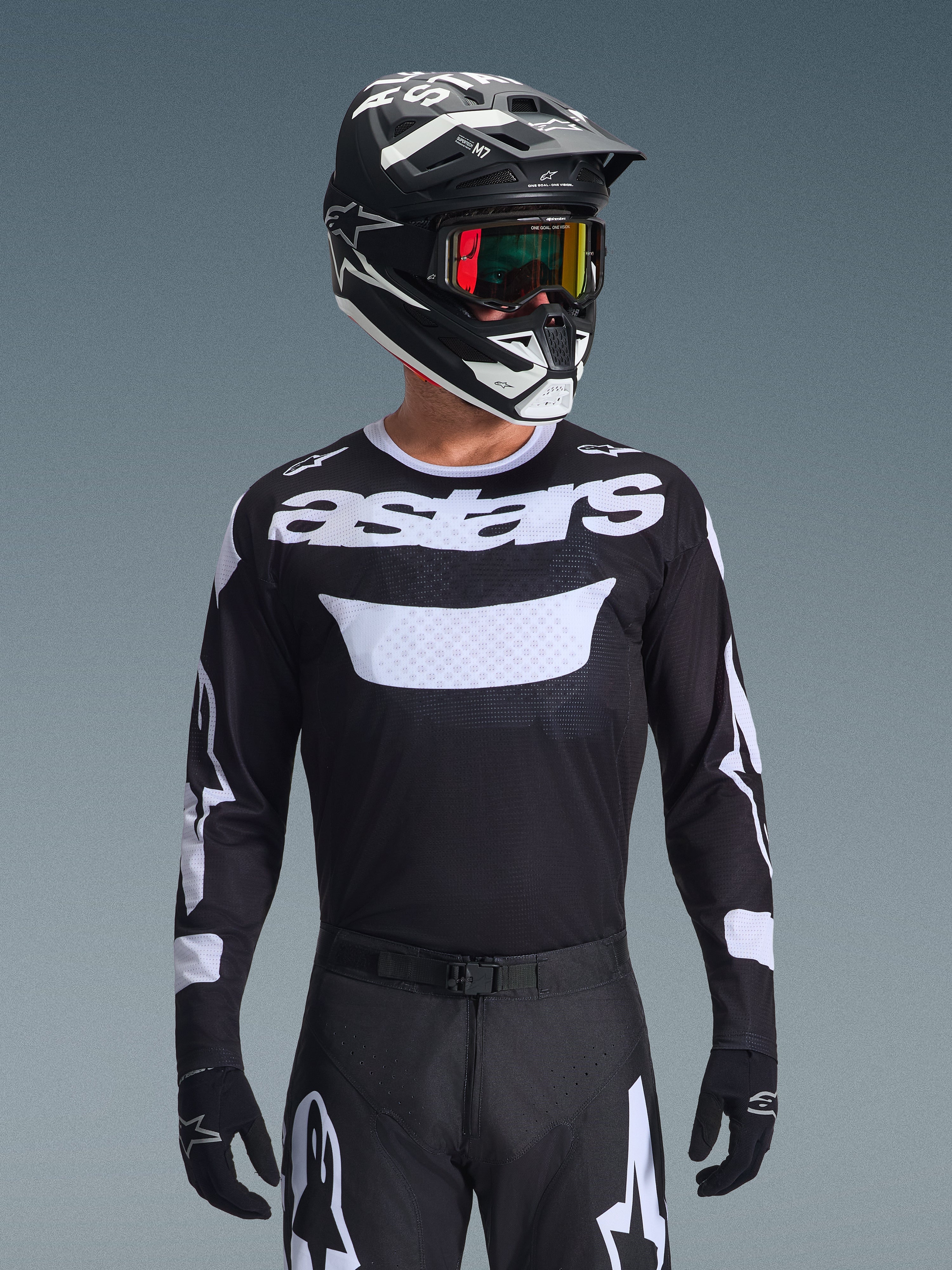 Pilota di motocross che indossa il completo Alpinestars 2026 Racer Air Riway, maglia nera con logo 'astars' bianco e pannello ventilato sul petto, pantaloni neri con accenti bianchi, casco coordinato nero e bianco con occhiali iridescenti, guanti neri, abbigliamento da motocross ad alte prestazioni in nero e bianco.