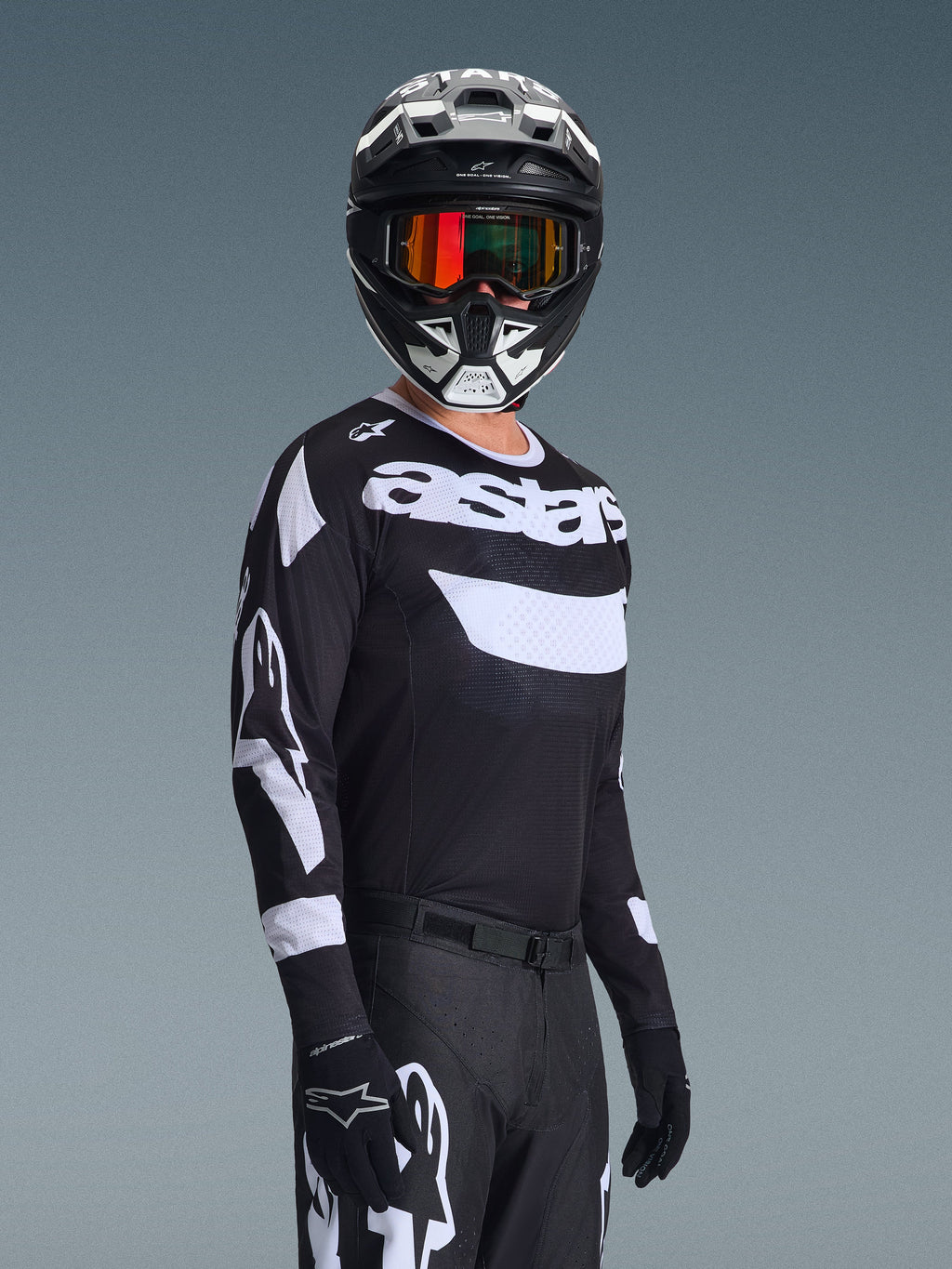 Pilota di motocross che indossa la maglia Alpinestars Racer Air Riway 2026, maglia bianca e nera con logo 'astars', casco coordinato bianco e nero con maschera a specchio arancione, pantaloni e guanti da motocross neri, outfit coordinato per gare off-road