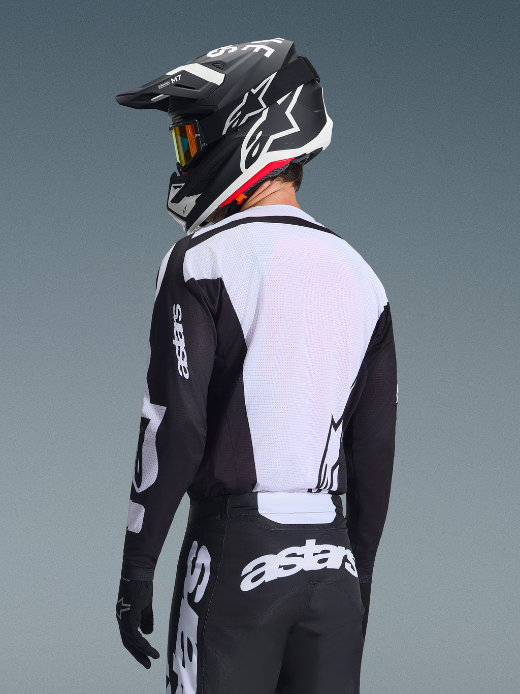 Pilota di motocross visto da dietro che indossa la maglia Alpinestars Racer Air Riway 2026, nera e bianca, con pannello posteriore in mesh bianco traspirante e maniche nere con marchio 'astars', pantaloni neri coordinati con loghi bianchi, casco nero con dettagli bianchi, abbigliamento off-road professionale per prestazioni ad alte temperature.