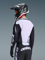 Pilota di motocross visto da dietro che indossa la maglia Alpinestars Racer Air Riway 2026, nera e bianca, con pannello posteriore in mesh bianco traspirante e maniche nere con marchio 'astars', pantaloni neri coordinati con loghi bianchi, casco nero con dettagli bianchi, abbigliamento off-road professionale per prestazioni ad alte temperature.
