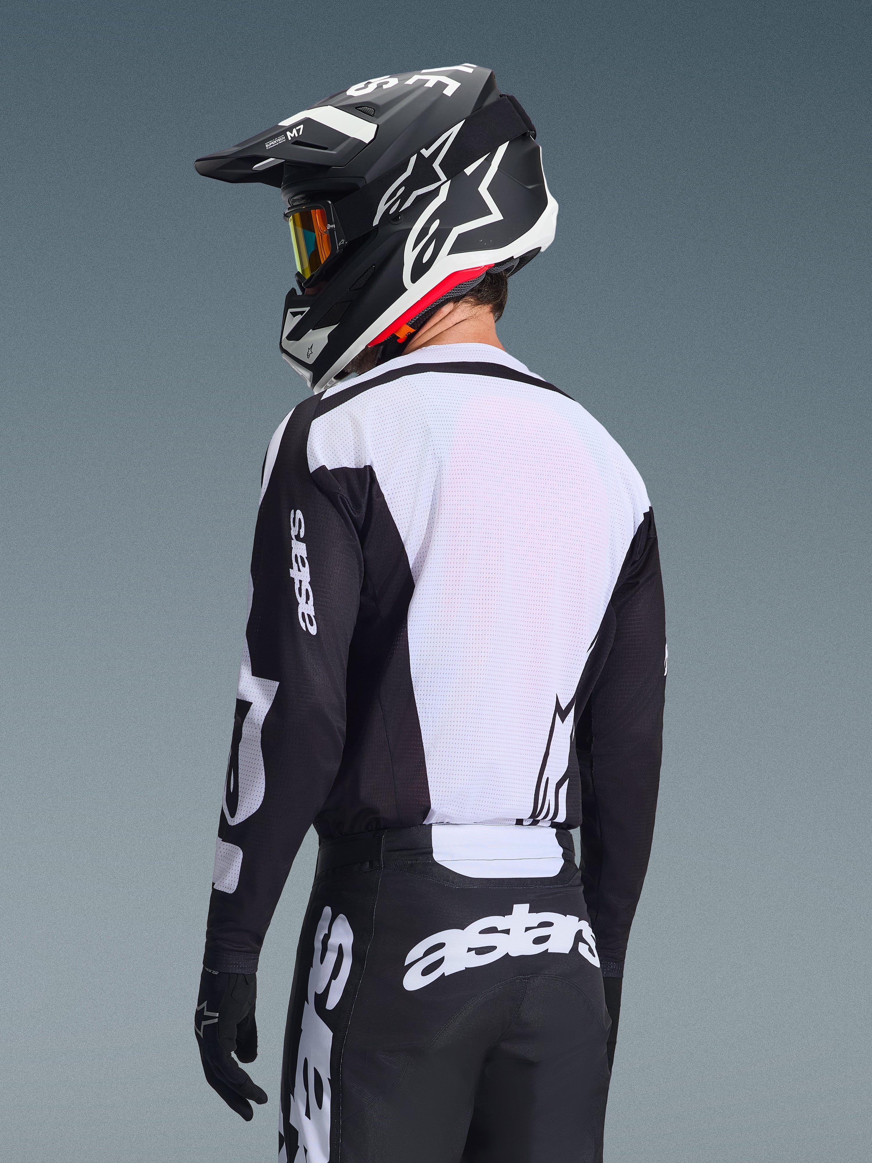 Pilota di motocross visto da dietro che indossa la maglia Alpinestars Racer Air Riway 2026, nera e bianca, con pannello posteriore in mesh bianco traspirante e maniche nere con marchio 'astars', pantaloni neri coordinati con loghi bianchi, casco nero con dettagli bianchi, abbigliamento off-road professionale per prestazioni ad alte temperature.