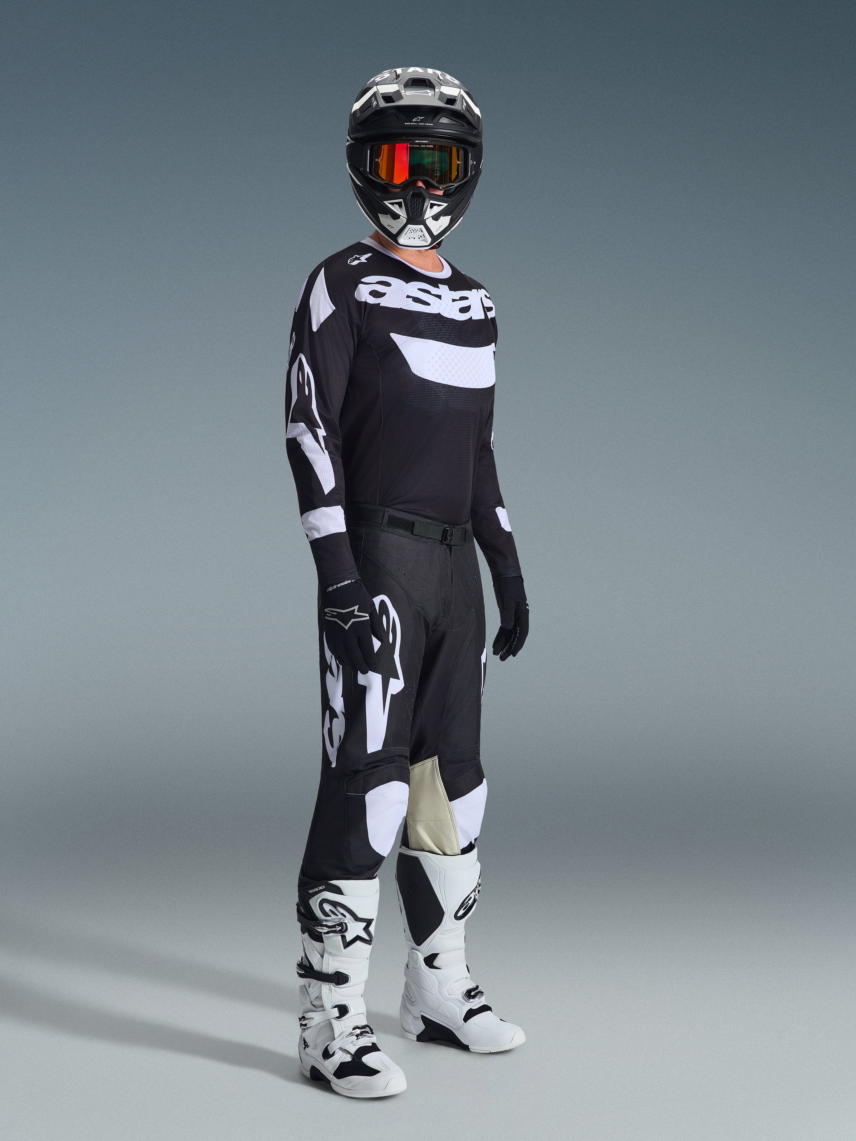 Pilota di motocross con abbigliamento completo da gara, maglia Racer Air Riway 2026 in nero e bianco, pantaloni neri coordinati con grandi loghi Alpinestars bianchi, casco nero con dettagli bianchi e occhiali arancioni, stivali bianchi e neri, completo coordinato per gare off-road professionali