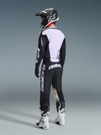 Pilota di motocross visto da dietro che indossa la maglia Racer Air Riway 2026 in bianco e nero, con l'ampio pannello posteriore in mesh bianco per favorire il flusso d'aria, abbinata a pantaloni neri, casco bianco e nero e stivali da gara bianchi, abbigliamento professionale completo per prestazioni off-road.