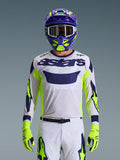 Maglia Alpinestars Racer Air Riway 2026, grigio chiaro con logo astars blu scuro, accenti giallo fluo e blu sulle maniche, indossata da un pilota di motocross con casco e guanti coordinati viola e giallo neon, tessuto tecnico in rete per gare off-road