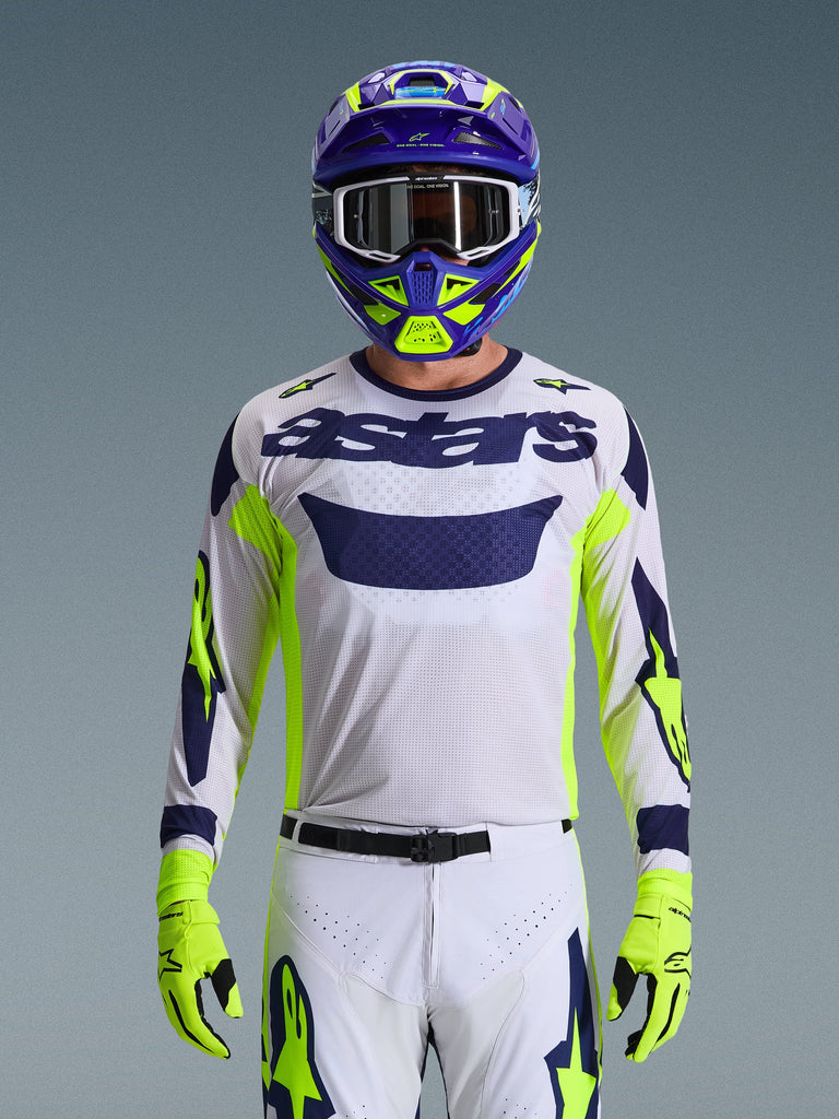 Maglia Alpinestars Racer Air Riway 2026, grigio chiaro con logo astars blu scuro, accenti giallo fluo e blu sulle maniche, indossata da un pilota di motocross con casco e guanti coordinati viola e giallo neon, tessuto tecnico in rete per gare off-road