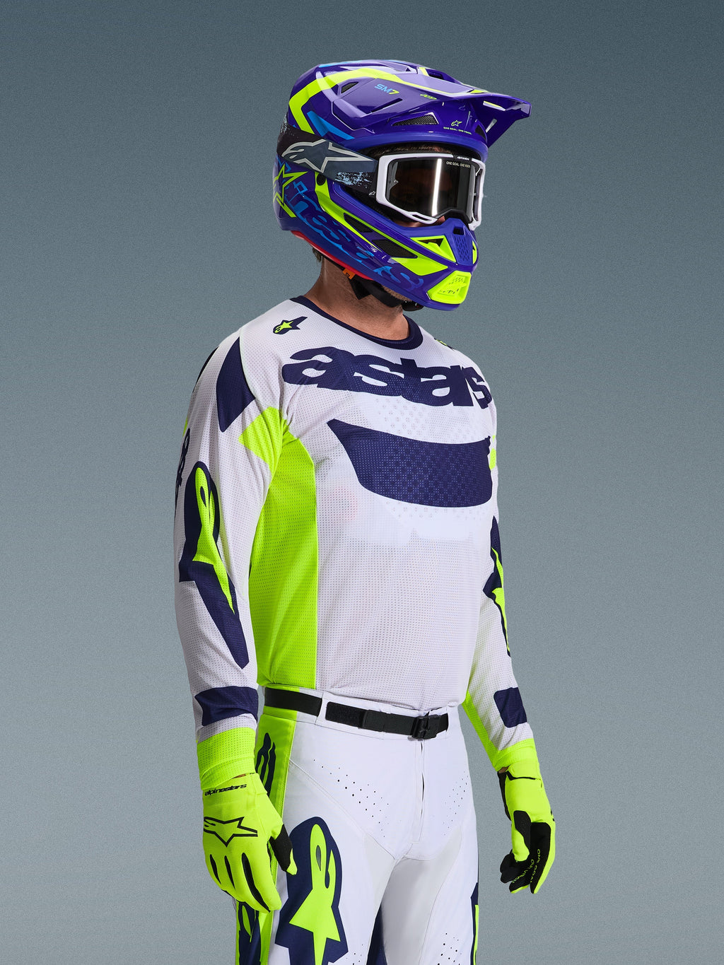 Pilota di motocross che indossa la maglia Alpinestars 2026 Racer Air Riway, grigio chiaro con accenti giallo fluo e blu, abbinata a un casco coordinato blu e giallo e guanti giallo fluo, con struttura in rete traspirante e stile racing professionale per prestazioni off-road.