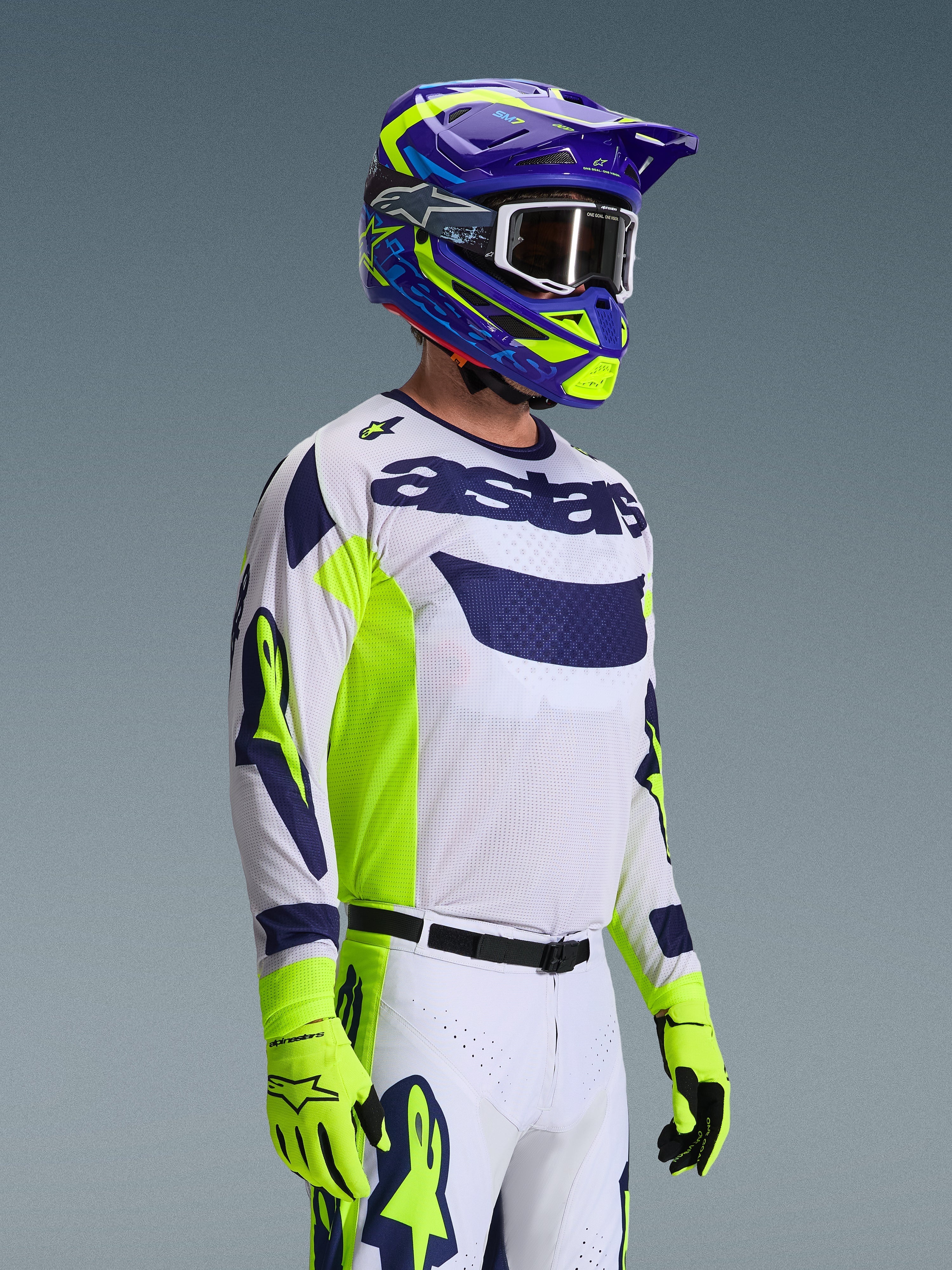 Pilota di motocross che indossa la maglia Alpinestars 2026 Racer Air Riway, grigio chiaro con accenti giallo fluo e blu, abbinata a un casco coordinato blu e giallo e guanti giallo fluo, con struttura in rete traspirante e stile racing professionale per prestazioni off-road.