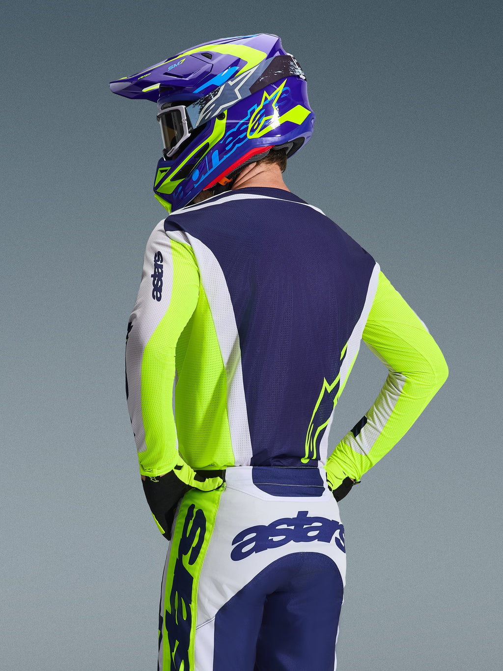 Alpinestars Racer Air Riway Jersey 2026, maglia da motocross, grigio chiaro, giallo fluo e blu, vista posteriore del pilota con pantaloni e casco coordinati, vestibilità atletica con pannelli in rete traspirante per corse off-road