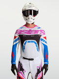 Alpinestars Maglia Supertech Dyna, maglia da motocross da uomo, bianca, blu e rosa, in mesh elasticizzato e traspirante, vestibilità racing per gare MX, indossata da un pilota con casco bianco e guanti neri.