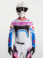 Alpinestars Maglia Supertech Dyna, maglia da motocross da uomo, bianca, blu e rosa, in mesh elasticizzato e traspirante, vestibilità racing per gare MX, indossata da un pilota con casco bianco e guanti neri.