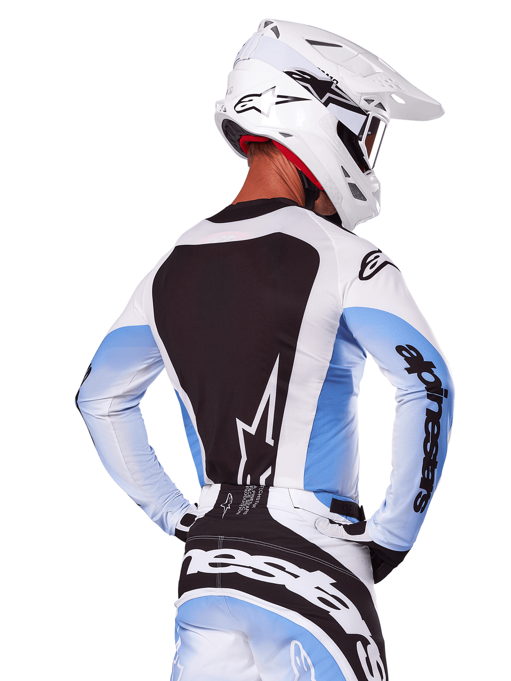 Vista posteriore di un pilota di motocross che indossa il set completo Alpinestars Techstar Melt 2025 in bianco e blu, maglia con ampio pannello posteriore nero e logo a stella bianco, maniche con sfumature azzurre, pantaloni coordinati bianchi e blu, casco da gara bianco e guanti bianchi e neri, abbigliamento tecnico per motocross
