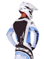 Vista posteriore di un pilota di motocross che indossa il set completo Alpinestars Techstar Melt 2025 in bianco e blu, maglia con ampio pannello posteriore nero e logo a stella bianco, maniche con sfumature azzurre, pantaloni coordinati bianchi e blu, casco da gara bianco e guanti bianchi e neri, abbigliamento tecnico per motocross