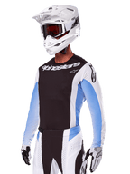 Pilota di motocross con Set Gear Alpinestars Techstar Melt 2025, bianco con dettagli neri e azzurri, maglia con petto nero e maniche bianche, pantaloni coordinati bianchi e neri, casco SM10 bianco con occhiali e guanti bianchi e neri, completo professionale per corse fuoristrada.