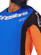 Alpinestars 2025 Techstar Melt Jersey, maglia da motocross, blu e arancione con pannelli neri, con marchio Alpinestars bianco e perforazioni laser visibili sul petto, indossata da un pilota con casco nero