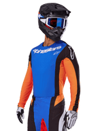Set completo da motocross Alpinestars Techstar Melt 2025, pilota con maglia blu, maniche arancioni e pannelli laterali neri, pantaloni coordinati blu e neri, casco nero con occhiali blu, guanti bianchi e neri, abbigliamento tecnico con vestibilità racing