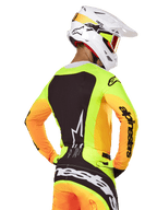 Pilota di motocross che indossa la maglia Alpinestars Techstar Melt 2025, vista posteriore, giallo neon con maniche arancioni e un grande pannello grafico nero, pantaloni da motocross coordinati con branding audace, casco Alpinestars bianco e maschera arancione, abbigliamento professionale per fuoristrada