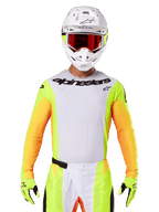 Alpinestars Techstar Melt Jersey 2025, abbigliamento da motocross, petto bianco con maniche giallo fluo e arancioni, pilota con casco bianco e maschera rossa, guanti giallo fluo con loghi neri, colorazione vivace per prestazioni off-road