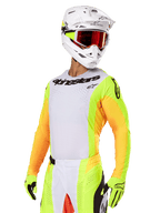 Pilota di motocross con set Alpinestars Techstar Melt 2025, maglia bianca con maniche giallo vivace e arancione, logo Alpinestars nero sul petto, guanti giallo neon coordinati, casco bianco con occhiali dalle lenti rosse, kit professionale da gara off-road