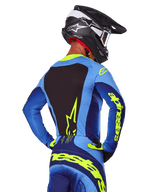 Vista posteriore di un pilota di motocross con maglia Alpinestars Techstar Melt 2025 blu e giallo fluo, pannello posteriore nero e logo giallo sulle maniche, abbinata a pantaloni coordinati e casco in carbonio nero, abbigliamento tecnico da gara off-road