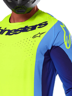 Maglia Alpinestars Techstar Melt 2025, maglia da motocross, giallo fluo e blu, con perforazioni tagliate al laser e logo vistoso sul petto, vestibilità atletica per gare off-road, vista ravvicinata con casco nero