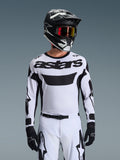 Pilota di motocross che indossa la maglia Alpinestars 2026 Racer Riway in bianco e nero, con logo 'astars' sul petto, abbinata a pantaloni coordinati bianco e nero, casco nero con occhiali e guanti neri, kit coordinato per corse off-road