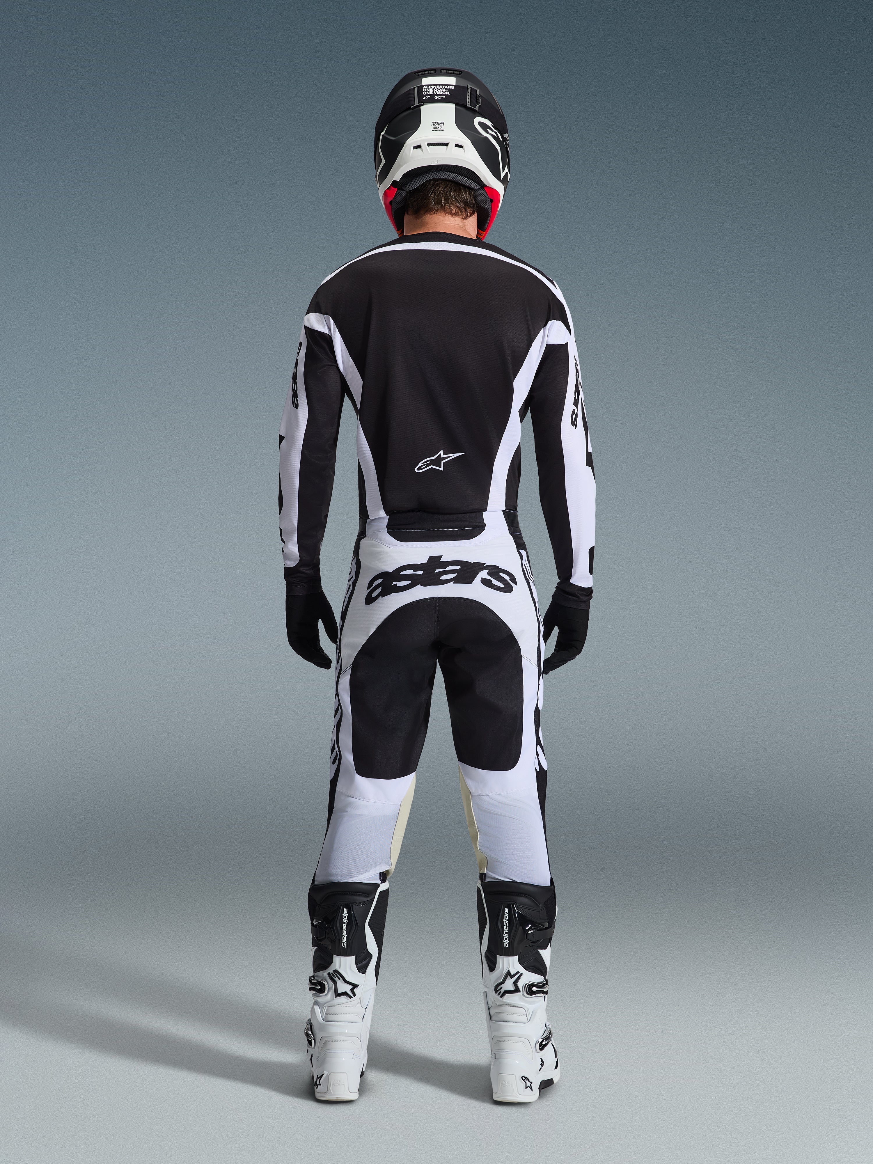 Pilota di motocross visto da dietro con abbigliamento da gara completo, maglia Racer Riway 2026 bianca e nera, pantaloni coordinati con marchio 'astars' sulla seduta, casco nero con dettagli bianchi, guanti neri e stivali bianchi e neri, outfit professionale coordinato per il fuoristrada