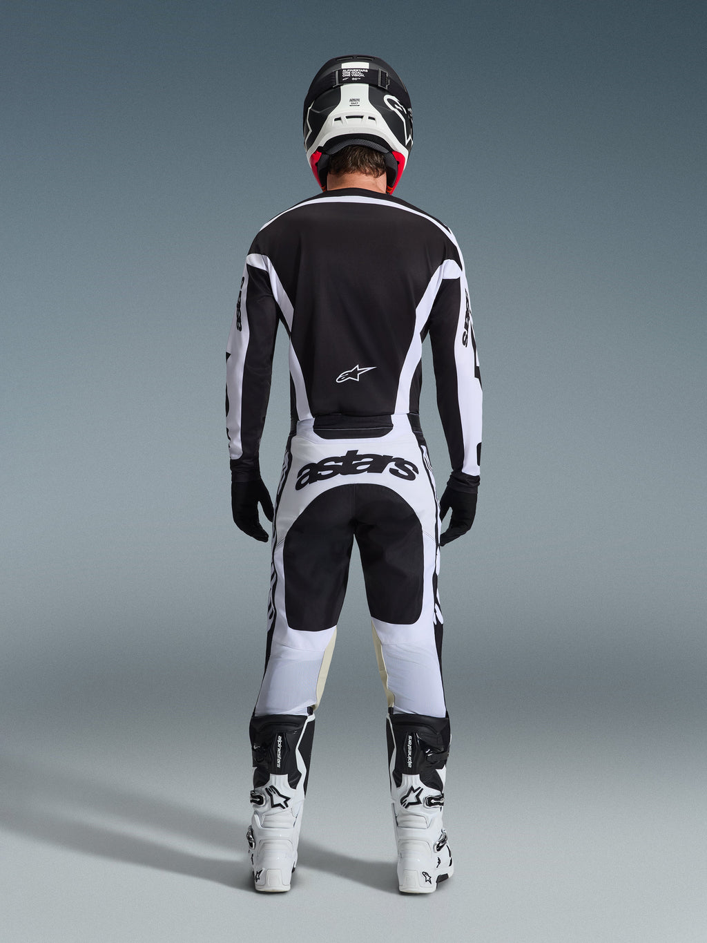 Vista posteriore di un pilota di motocross con il completo 2026 Racer Riway in bianco e nero, caratterizzato da maglia nera con accenti bianchi, pantaloni bianchi con vistoso marchio 'astars' sulla seduta, casco nero, stivali bianchi e guanti neri, abbigliamento professionale completo da gara off-road.