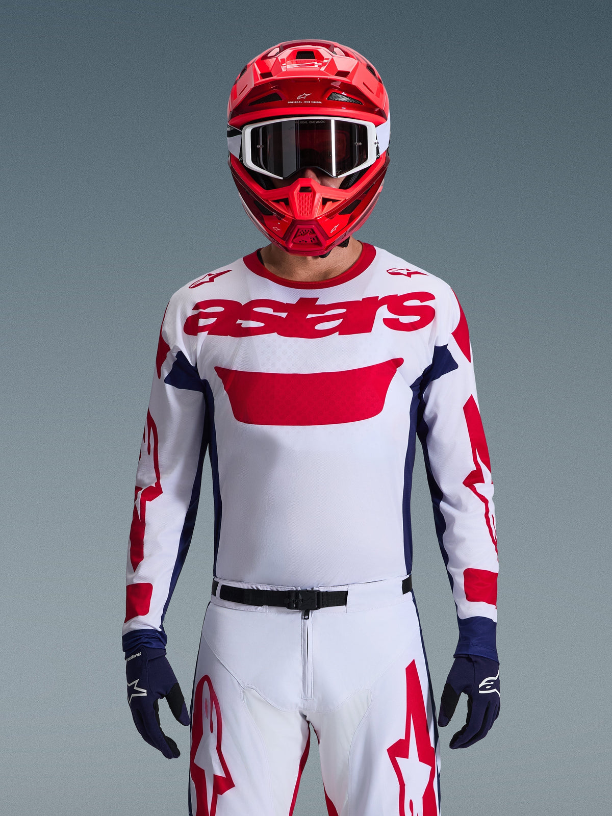 Pilota di motocross che indossa la maglia Alpinestars 2026 Racer Riway in White Blue Red, caratterizzata da un grande logo 'astars' rosso sul petto, pannelli laterali blu navy, pantaloni da motocross coordinati bianchi e rossi, casco rosso con occhiali bianchi e guanti blu navy, set completo da gara off-road.
