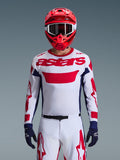 Pilota di motocross che indossa la maglia Alpinestars 2026 Racer Riway in White Blue Red, caratterizzata da un grande logo 'astars' rosso sul petto, pannelli laterali blu navy, pantaloni da motocross coordinati bianchi e rossi, casco rosso con occhiali bianchi e guanti blu navy, set completo da gara off-road.