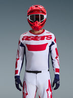 Pilota di motocross che indossa la maglia Alpinestars 2026 Racer Riway in White Blue Red, caratterizzata da un grande logo 'astars' rosso sul petto, pannelli laterali blu navy, pantaloni da motocross coordinati bianchi e rossi, casco rosso con occhiali bianchi e guanti blu navy, set completo da gara off-road.